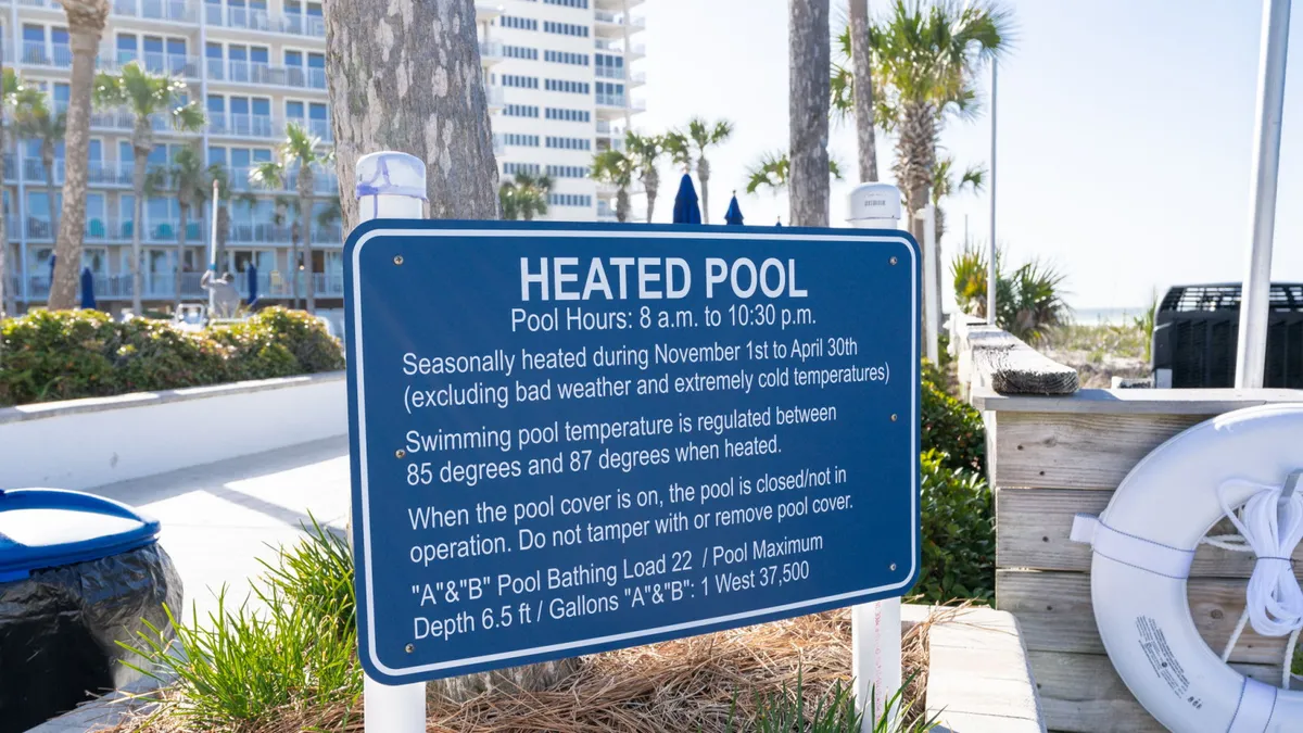 A bright exterior view of the heated pool area, complete with surrounding palm trees, a clearly marked 'Heated Pool' sign, a pool deck with lounge chairs, a lifebuoy, and a pool cover. The atmosphere is cheerful, suggesting year-round swimming comfort even in cooler weather.