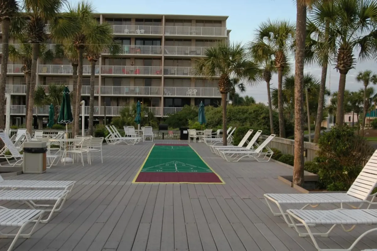 A lively poolside scene featuring lounge chairs, a shuffleboard court, palm trees, and balconies overlooking the outdoor amenities. People can be seen engaging in games and relaxing, embodying a playful family atmosphere.