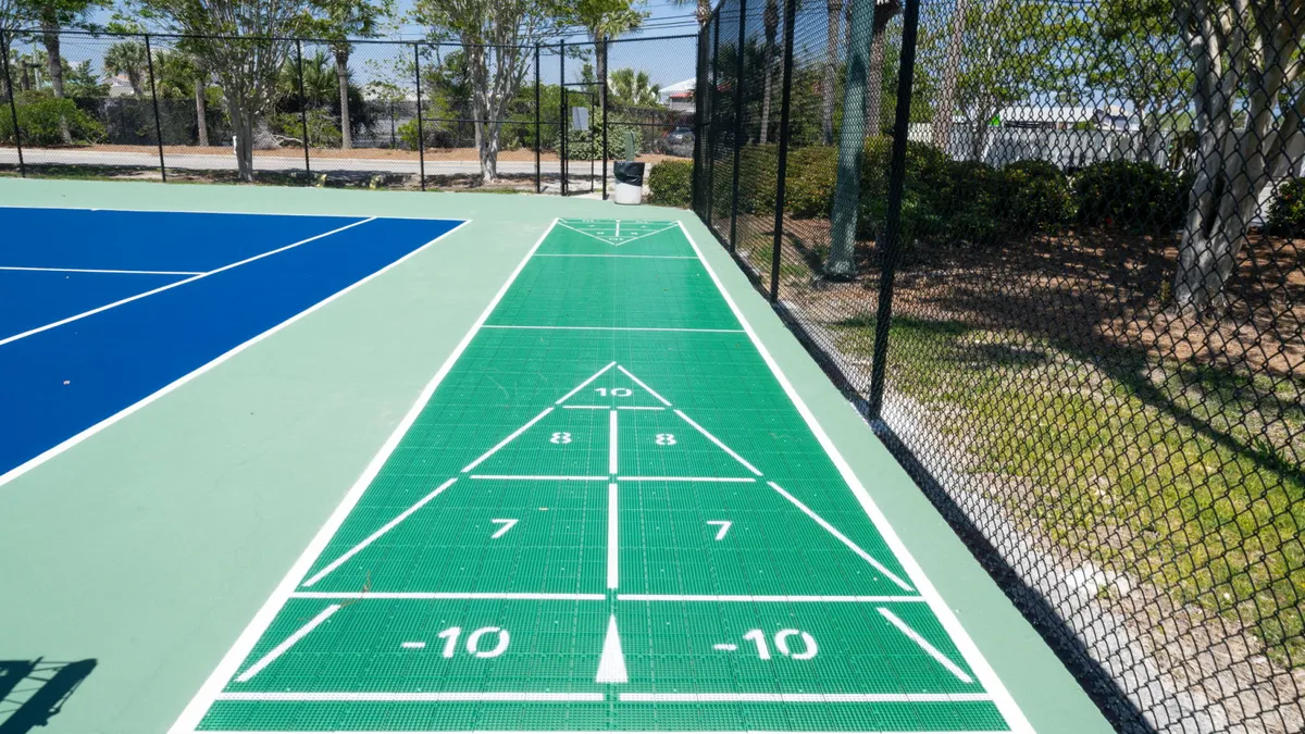 A vibrant exterior scene featuring both tennis and shuffleboard courts, set against a backdrop of greenery and blue skies. The courts are well-maintained and clearly designed for active, social play.