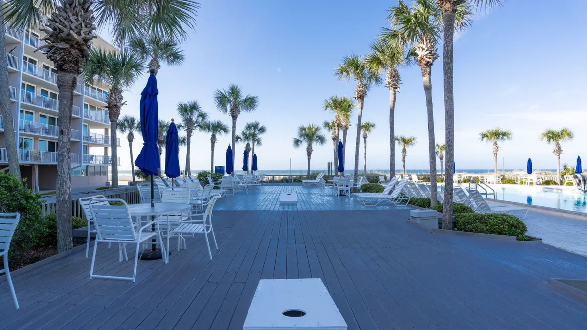 A sun-drenched swimming pool is surrounded by palm trees, blue umbrellas, and a comfortable seating area. The inviting pool area is designed for socializing and relaxation, with ample space for guests to lounge, swim, or gather for community events.