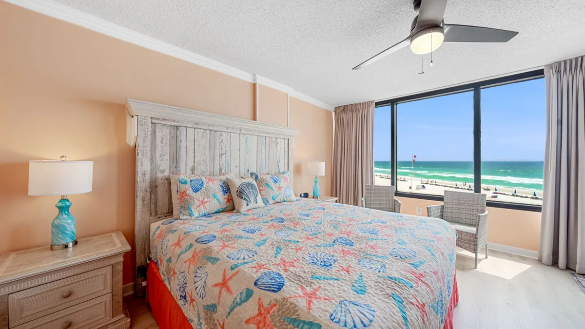 This image features a cozy bed with a colorful quilt positioned near large windows overlooking the ocean. A nightstand with a lamp adds to the bedroom's inviting feel, and the natural light flooding in enhances the serene, coastal ambiance.