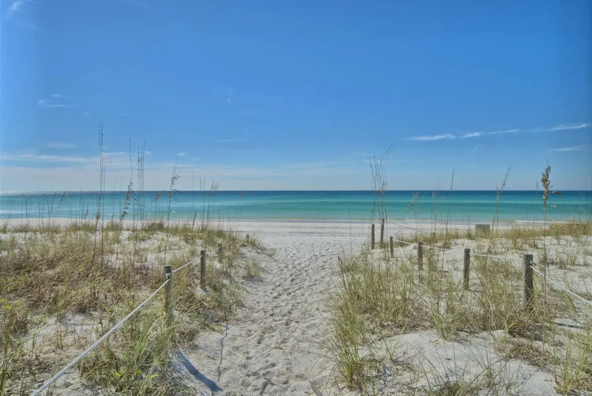 A serene view of a sandy pathway leading directly to the beach, bordered by gentle waves and a clear blue sky. The scene evokes tranquility, the promise of morning walks, and direct connection to the shore just steps from the resort.