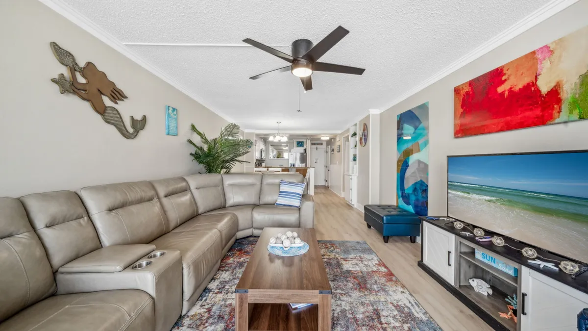 The image showcases a bright, open-concept living area featuring a spacious L-shaped leather sectional sofa, a wall-mounted flat-screen TV displaying a beach scene, colorful abstract artwork, and a modern wooden coffee table with a decorative bowl. The inviting space flows seamlessly into a fully equipped kitchen and hints at the private balcony beyond, offering a glimpse of coastal living.