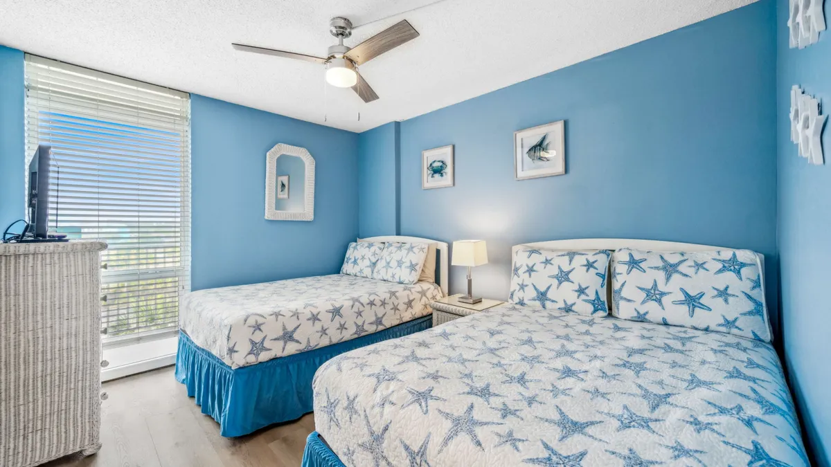The second bedroom is shown with two beds dressed in starfish patterned bedding, a blue accent wall, a window letting in natural light, and a white wicker dresser with a TV. The room is airy and inviting, making it perfect for families or groups seeking comfort and privacy.