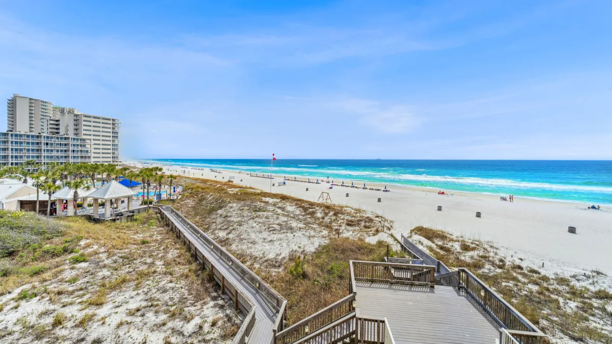 A scenic view of a wooden boardwalk leading over sand dunes to a wide, white sandy beach with scattered beachgoers. The ocean stretches to the horizon, and a large beachfront condominium building is visible, setting a classic Panama City Beach scene.