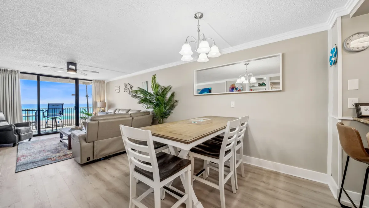 A spacious, sunlit living area with leather couches and a dining table for four, situated beside large glass sliding doors that open up to a private balcony. The ocean view from the balcony is clearly visible, and an indoor plant adds a touch of greenery and vibrancy to the space.