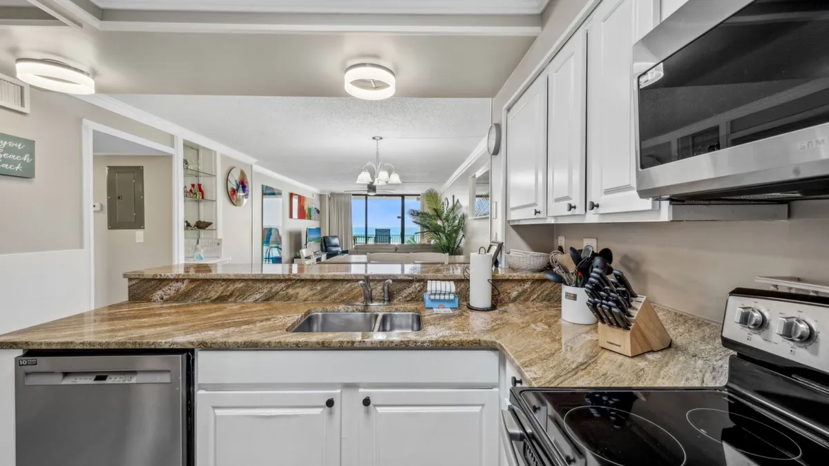 This image provides a broad view of the open-concept kitchen, dining area, and living room. The scene features a granite kitchen countertop with a modern sink, stainless steel appliances, a stylish chandelier above the dining area, and a living space that flows out to a balcony with sparkling ocean views.
