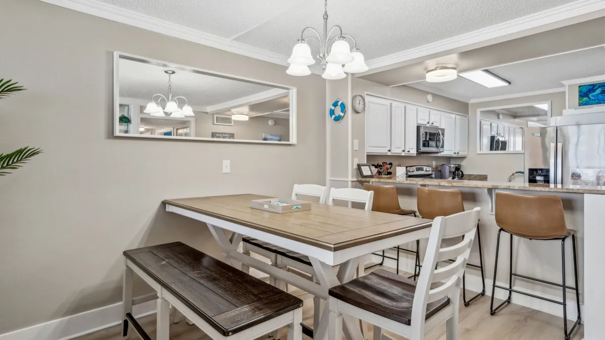 The image focuses on a welcoming dining setup, showcasing a wooden dining table with bench and chairs, a kitchen counter with bar stools, stainless steel appliances, and a large wall mirror that adds depth and brightness to the space.