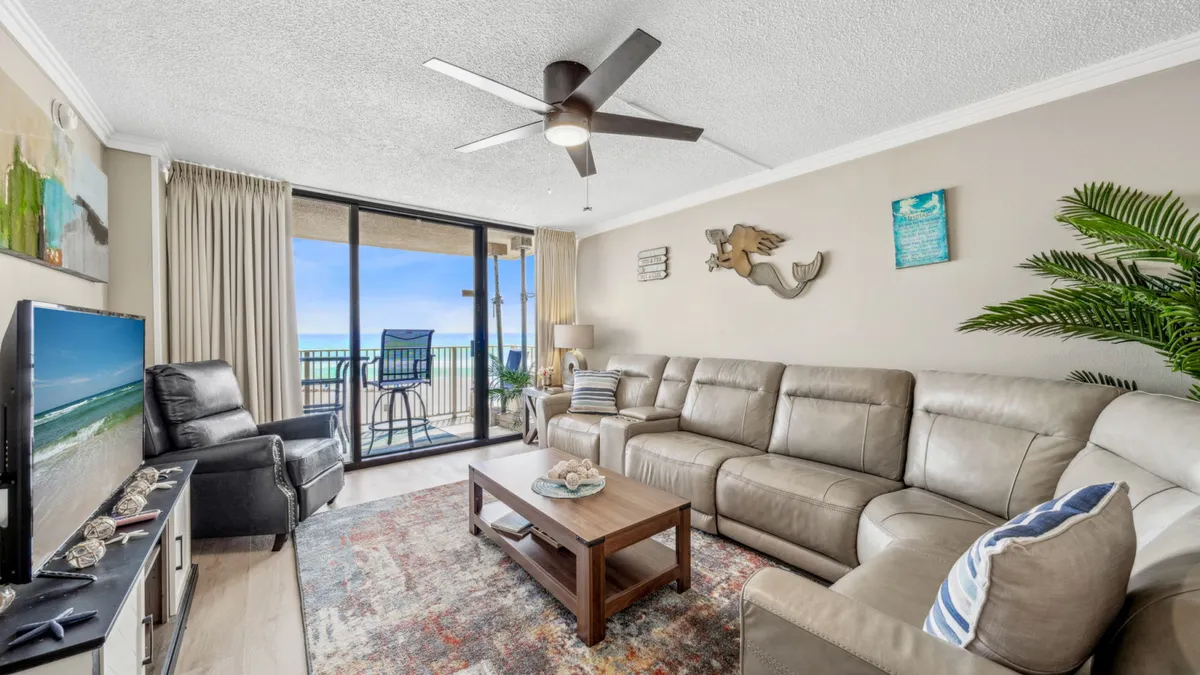 A large, comfortable leather sectional sofa faces both a balcony with sweeping ocean views and a flat-screen TV. Coastal wall art decorates the space, and natural light pours in from the balcony doors, enhancing the tranquil, inviting mood.