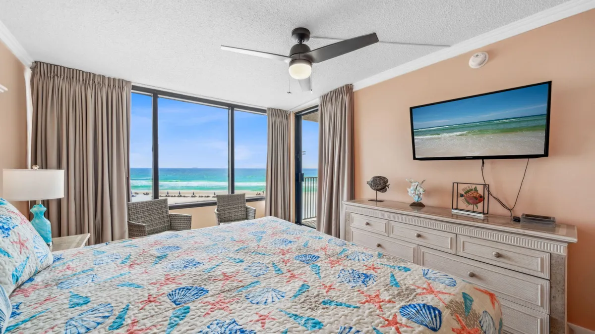 This image captures the primary suite, featuring a king-sized bed adorned with seashell patterned bedding. A large window frames a stunning beach and ocean view, and a mounted flat screen TV is visible above a dresser topped with coastal décor. The serene color palette and ample natural light highlight the room's restful, rejuvenating atmosphere.