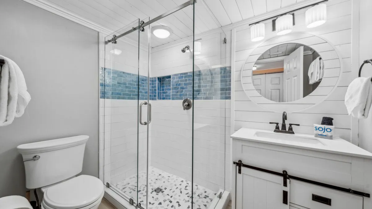 A bright, modern bathroom is featured with a glass-enclosed walk-in shower accented by blue tiles, a white vanity with round mirror, and a neatly arranged towel rack. The space feels clean, fresh, and spa-like, inviting guests to unwind after a day at the beach.