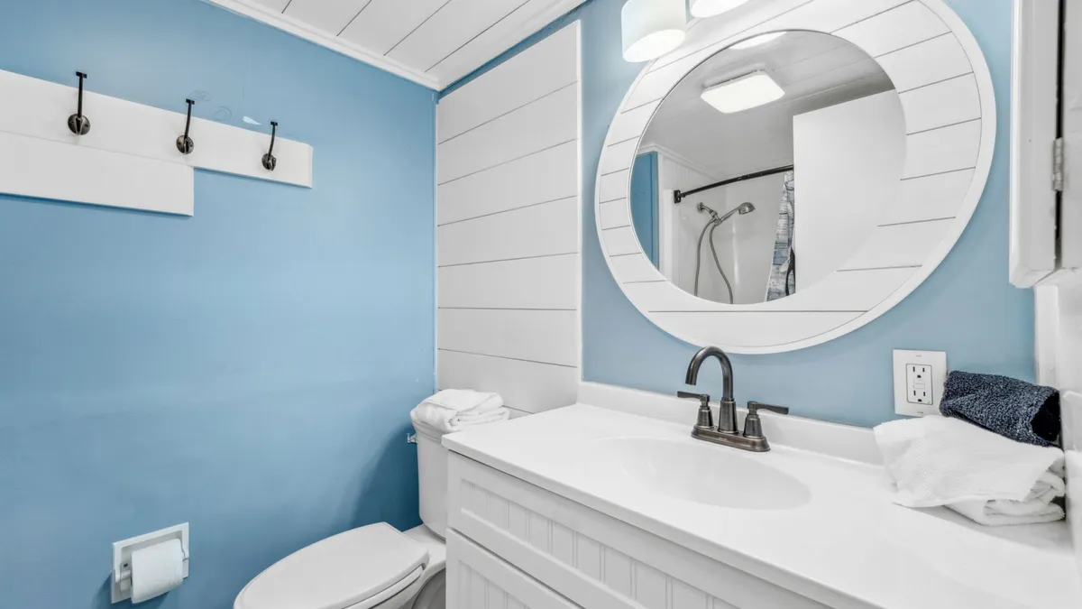 This bathroom features a white vanity with a bronze faucet, a round mirror, a toilet with neatly folded towels, and blue walls with white wainscoting. The space is bright, welcoming, and thoughtfully decorated with a coastal flair.