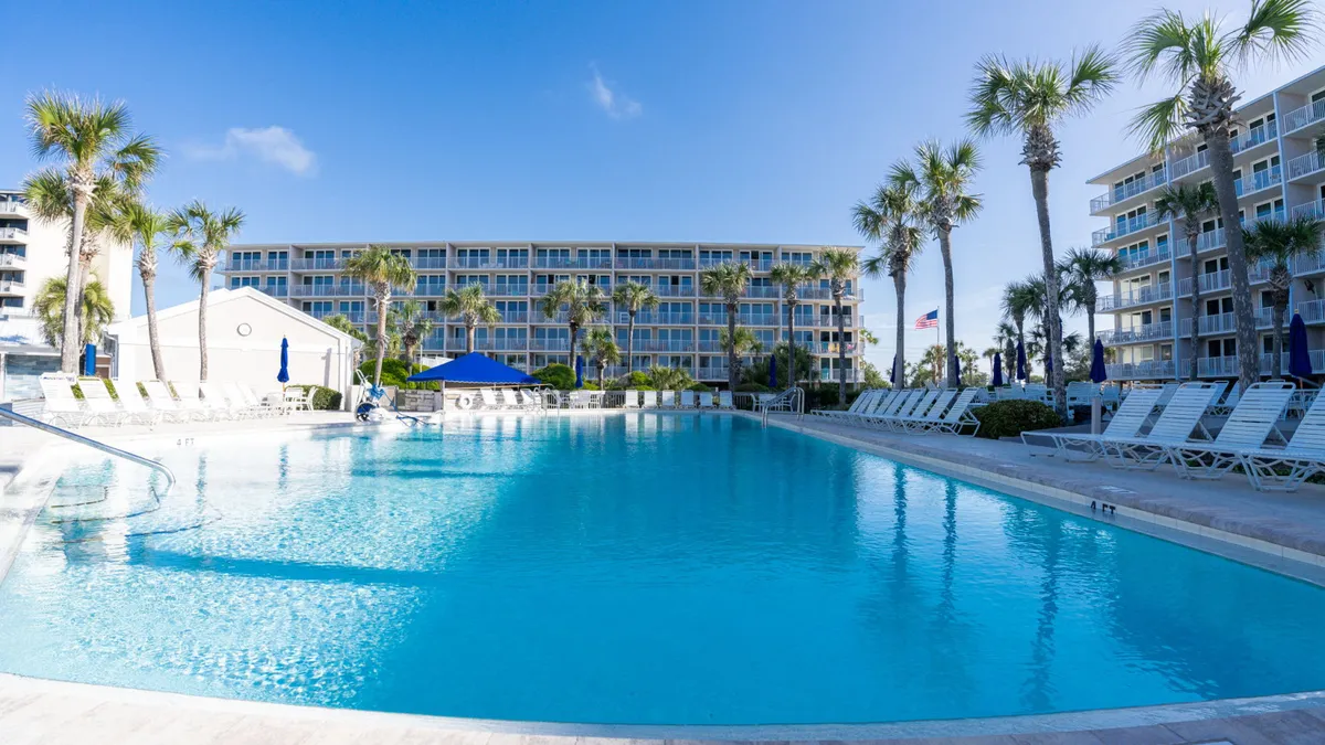 A large community swimming pool with clear blue water is surrounded by white lounge chairs, tall palm trees, and resort buildings in the background. Guests are swimming, lounging, and socializing in a tranquil, sun-filled setting.