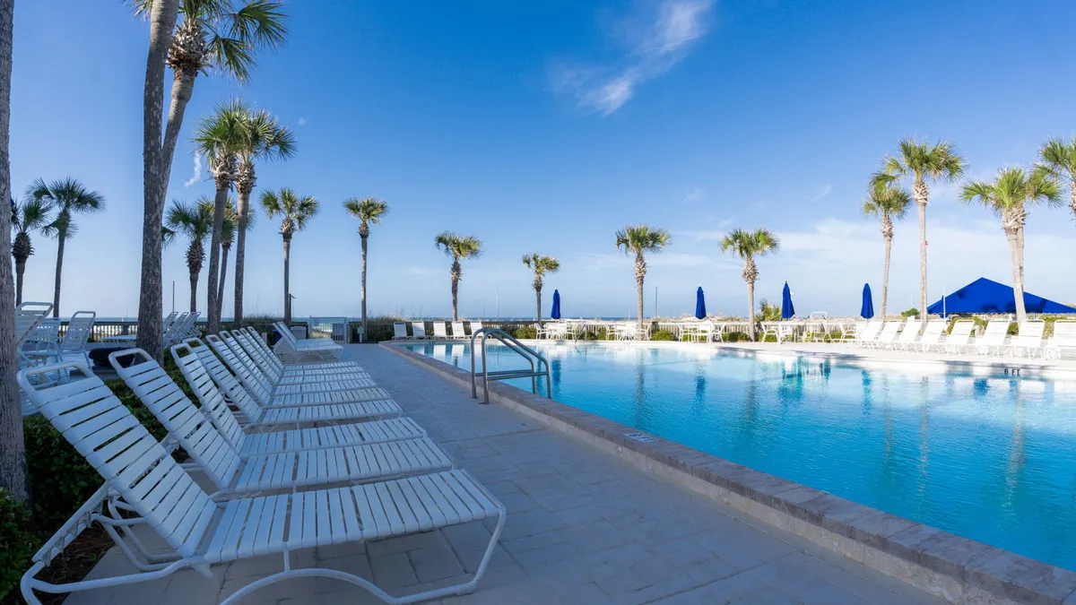 A large outdoor swimming pool with clear blue water is surrounded by neatly arranged white poolside lounge chairs, tall swaying palm trees, and blue umbrellas providing shaded seating. The pool area is tranquil and inviting, with views of the ocean in the background, suggesting a perfect spot for relaxation or socializing during the winter months.