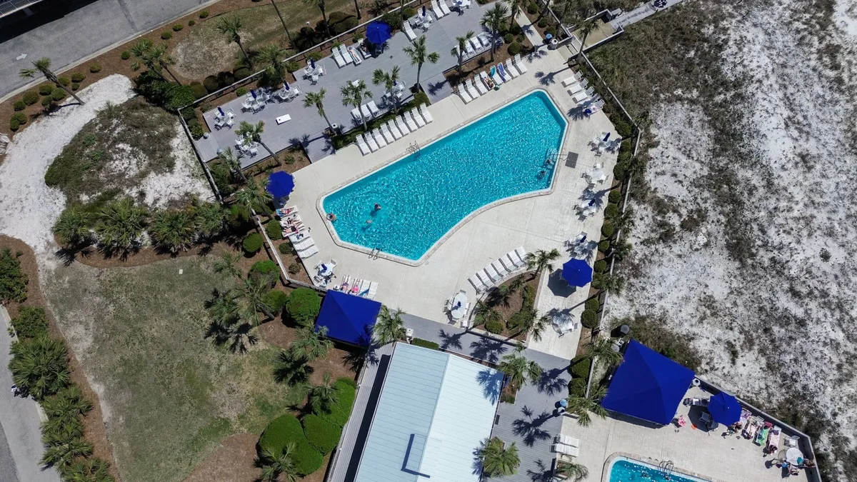A large community swimming pool with crystal-clear blue water is surrounded by neatly arranged poolside lounge chairs and blue umbrellas, offering plenty of shaded seating. Lush landscaping and sand dunes frame the area, reinforcing the resort's proximity to the beach.