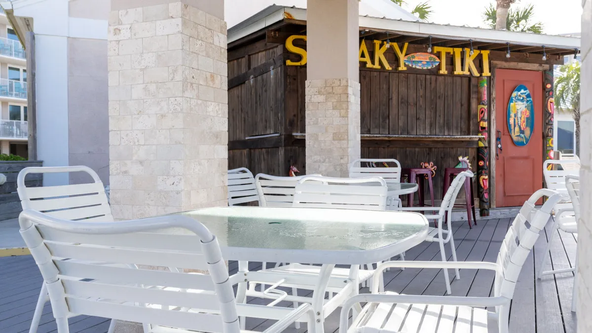 An inviting outdoor area featuring white dining chairs and a glass-top table is set near a charming wooden tiki bar with colorful painted doors and signage. The space is casual and lively, ideal for socializing, enjoying drinks, or dining in a scenic beachfront environment.