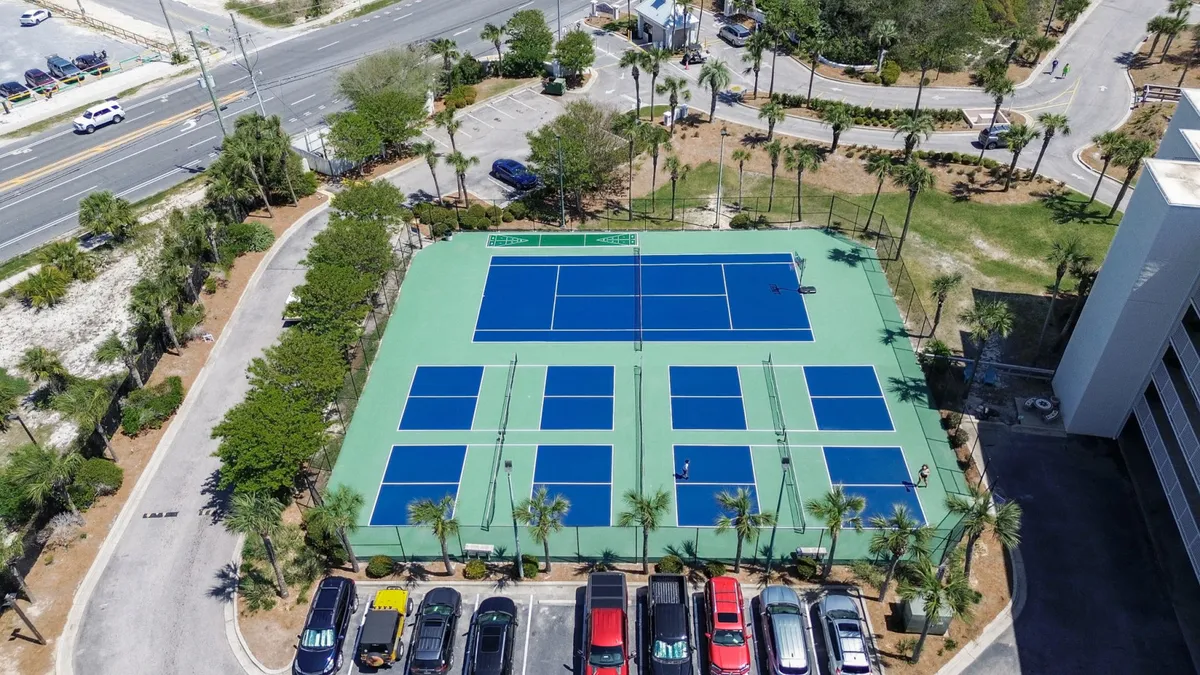This exterior image features the resort's tennis and pickleball courts surrounded by palm trees, with the parking lot and lush greenery visible in the background. The courts are clean, inviting, and clearly marked, providing a vibrant setting for active recreation.