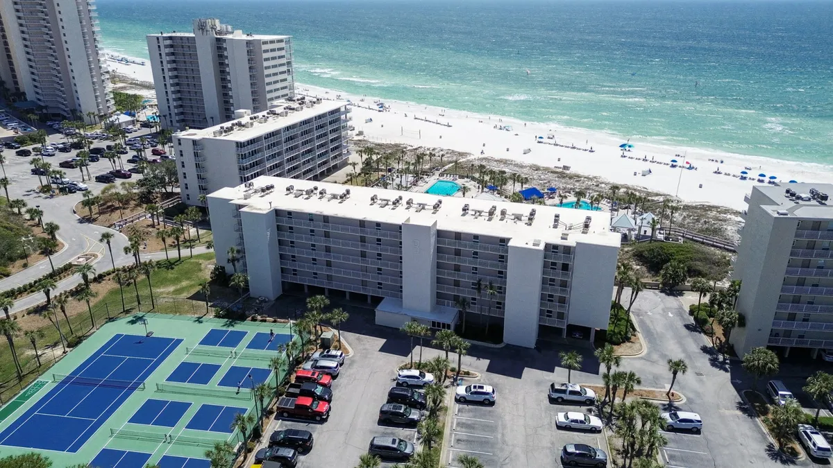 This wide-angle exterior shot features the Dunes of Panama building with balconies facing the beach, community tennis and pickleball courts, the white sandy beach with turquoise waves, and a parking lot lined with palm trees. The scene is lively with guests enjoying both sports and relaxation.
