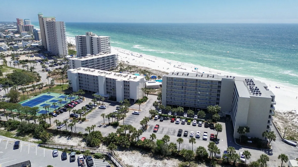 A beachfront condominium complex with private balconies faces a wide stretch of white sandy beach and turquoise ocean waters. Adjacent to the property are community tennis and pickleball courts, while palm trees and lush landscaping create a tropical, welcoming environment.