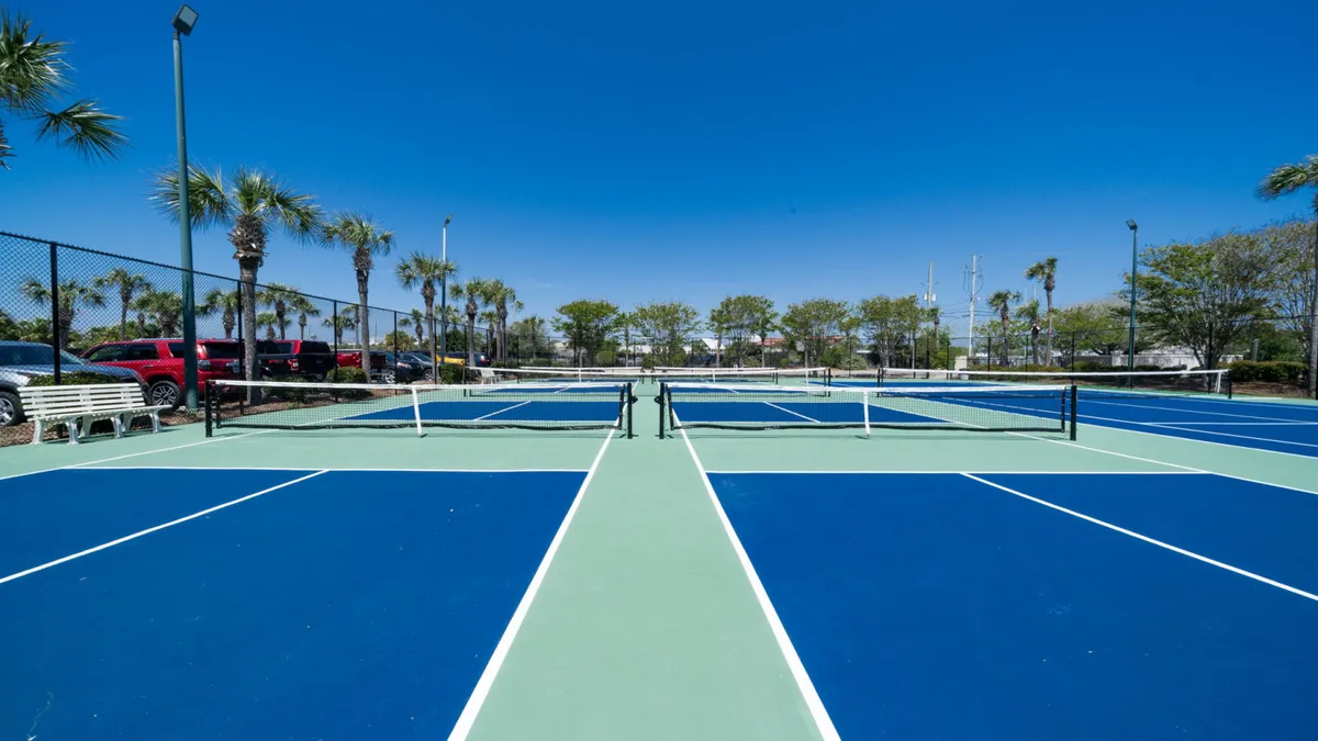 Outdoor pickleball courts with bright, clear markings and nets, surrounded by palm trees and benches for spectators or resting players. The blue sky and greenery create an energetic, inviting setting for outdoor sports.