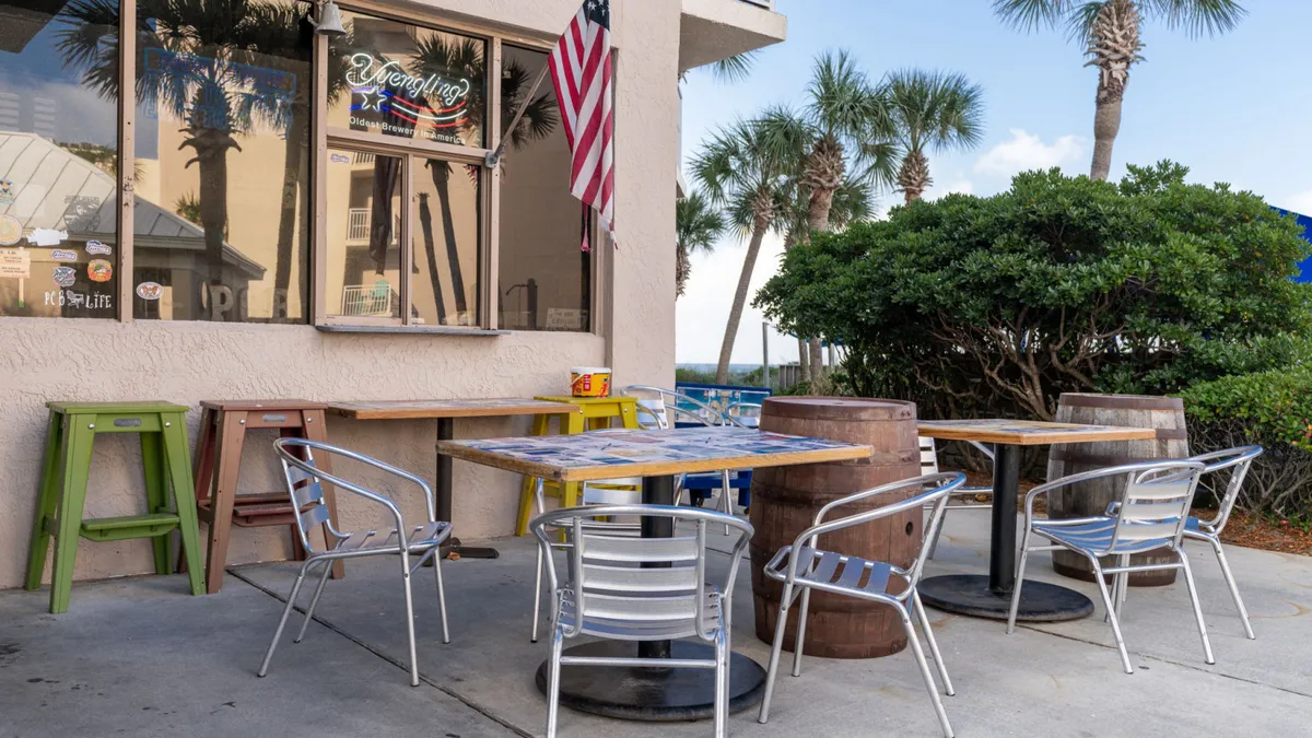 An outdoor seating area with metal chairs, wooden barrel tables, an American flag, and lush palm trees creates a laid-back, sociable vibe. The setting is perfect for al fresco dining or enjoying drinks, echoing the atmosphere of local eateries and bars.