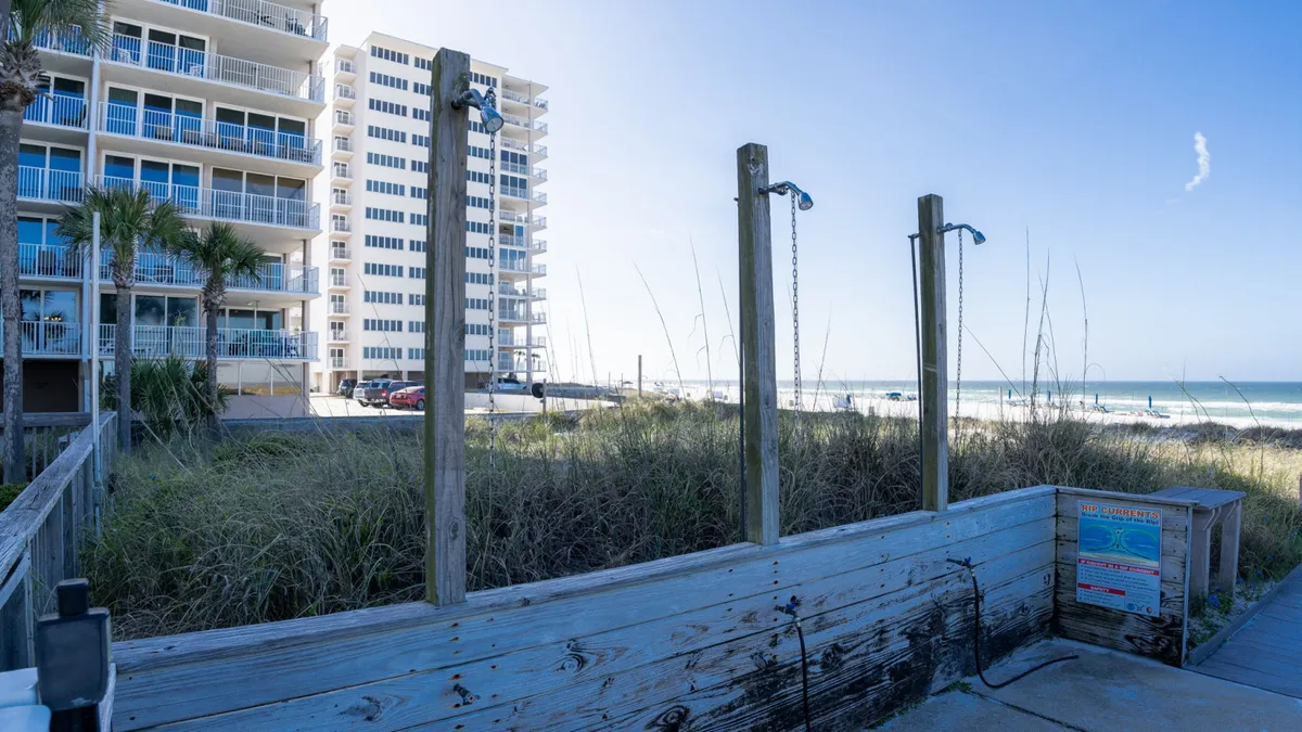 This exterior image showcases the beach showers with a weathered wood structure, the beachfront condo building with balconies overlooking the sand, ocean waves, and lush dune vegetation. Guests are seen rinsing off after swimming, walking along the beach, and relaxing on their balconies.