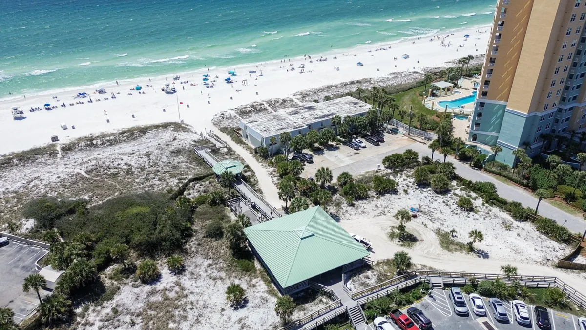 A lively, wide-angle beach scene with sunbathers and umbrellas dotting the pristine white sand, turquoise Gulf waters, a beachfront pavilion, and the resort building with its adjacent pool visible. The scene buzzes with leisurely vacation energy, highlighting both private and communal spaces.