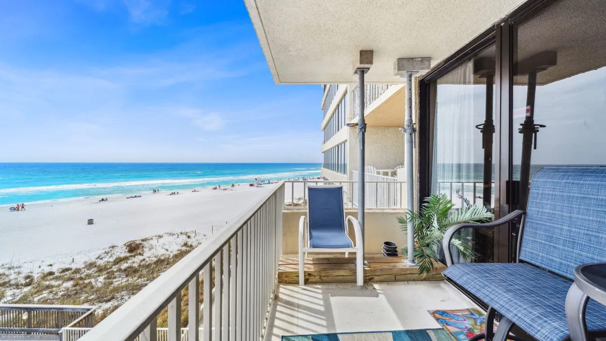 A private balcony furnished with blue cushioned chairs overlooks a pristine white sandy beach and the turquoise ocean beyond. The scene is tranquil and rejuvenating, ideal for sipping morning coffee or relaxing at sunset.